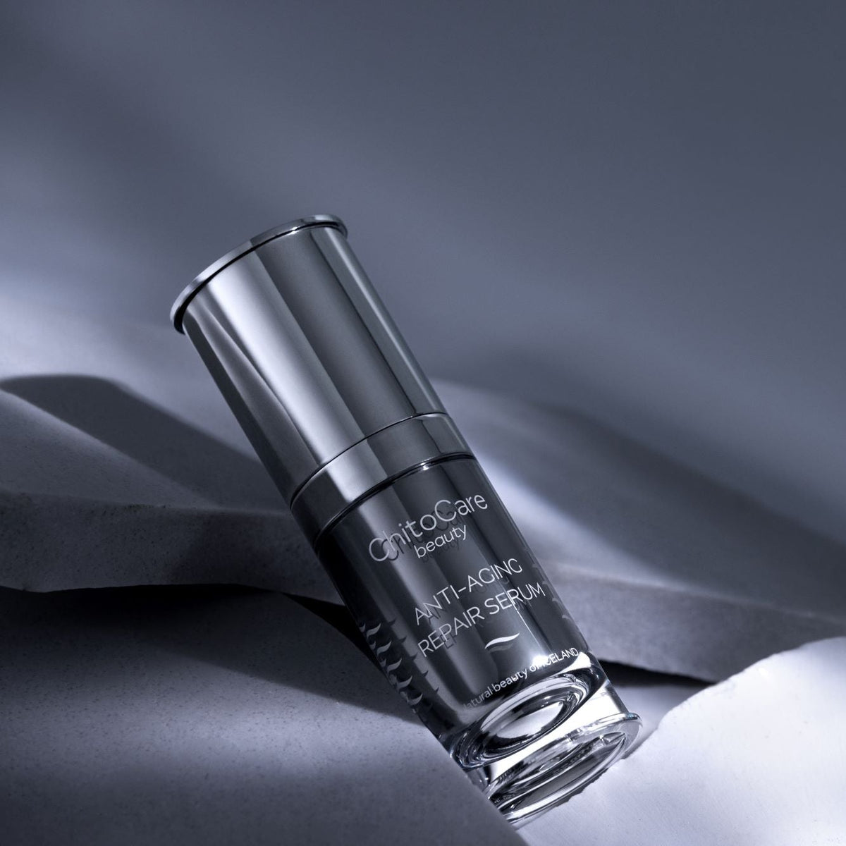 A bottle of ChitoCare Beauty Anti-Aging Repair Serum, featuring a sleek black and silver design, resting on a draped dark gray fabric background.