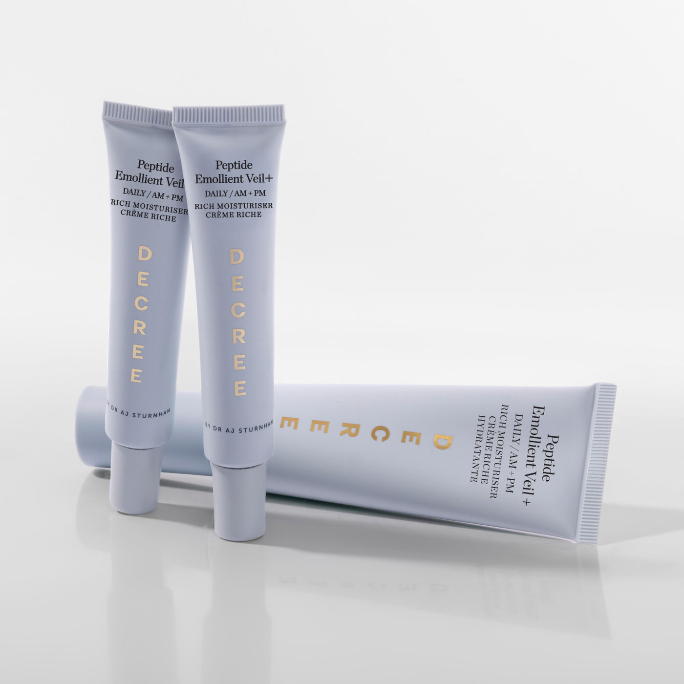 Decree Peptide Emollient Veil+ 50 ml packaging for dry, dehydrated, and fatigued-looking skin. Three Decree Enrichment Cream tubes in white and light gray packaging with gold lettering, arranged on a reflective white surface against a clean white background.