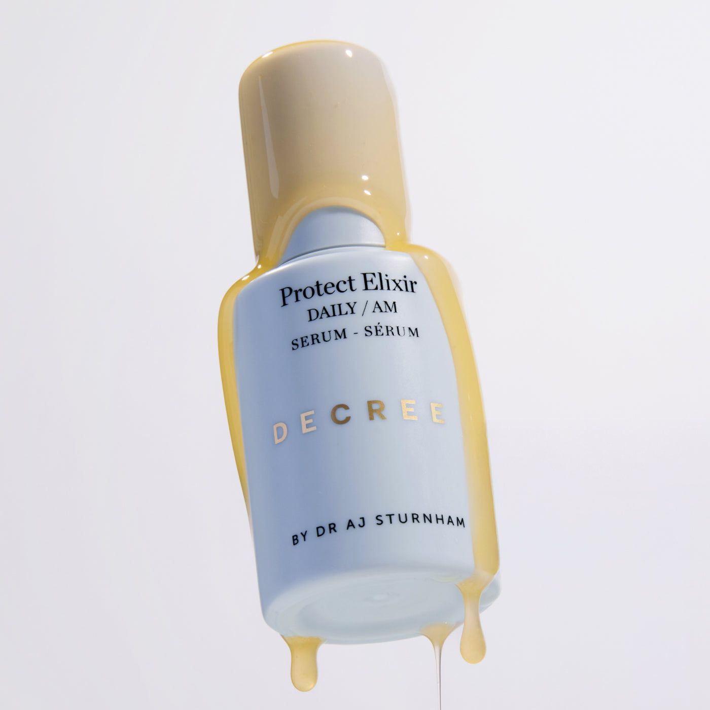 A bottle of Decree Protect Elixir serum, featuring a pale blue body with gold lettering and a beige cap, with golden liquid dripping from the top and bottom against a clean white background. Decree Protect Elixir serum texture with ascorbyl glucoside, vitamin E, ferulic acid, and coenzyme Q10.