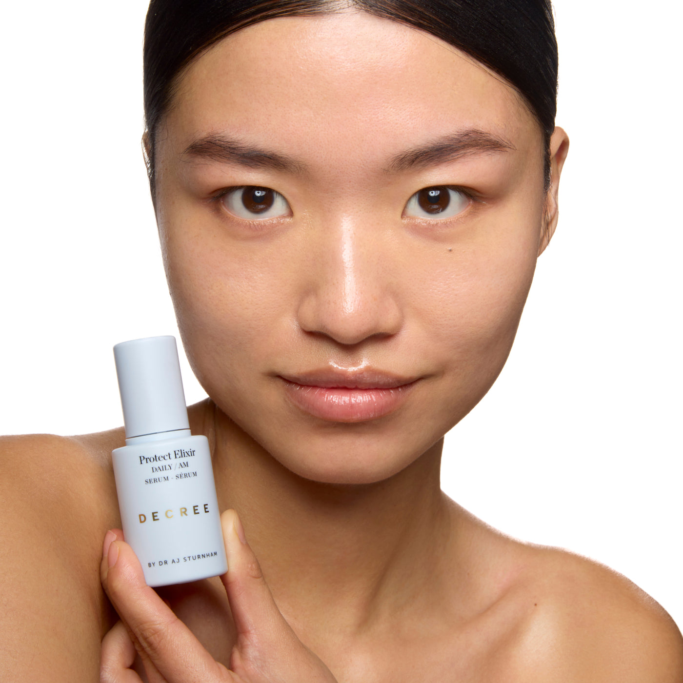 Woman holding a skincare serum bottle with a white background. Decree Protect Elixir applied to the face and neck as part of a morning skincare routine.