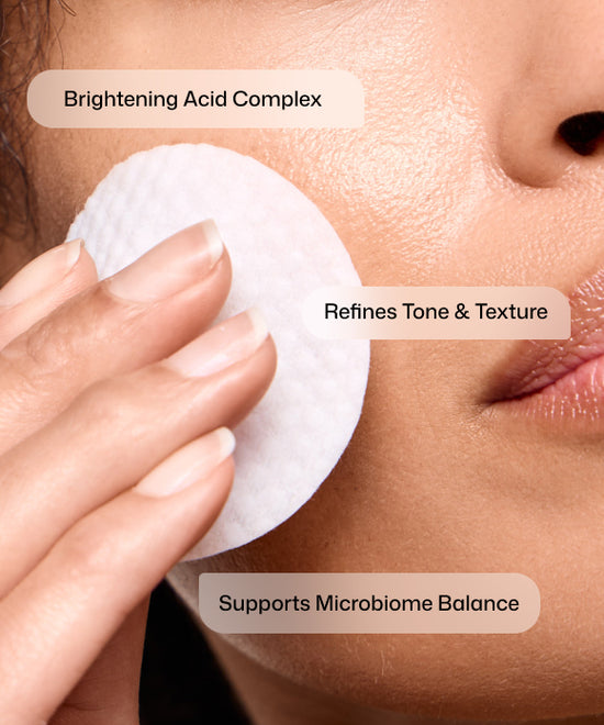Close-up of a person's face with a white round cotton pad applied to the cheek, overlaid with text reading 'Brightening Acid Complex,' 'Refines Tone & Texture,' and 'Supports Microbiome Balance.'. Revision Tone+ Correcting Pad applied to the face, neck, and décolletage during an evening skincare routine.