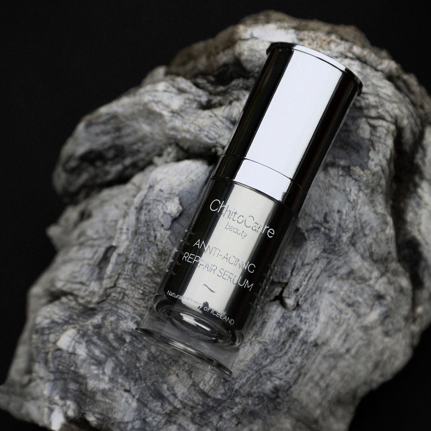 ChitoCare Anti-Aging Repair Serum in a clear bottle with a reflective silver cap, resting on a textured piece of weathered wood against a black background.