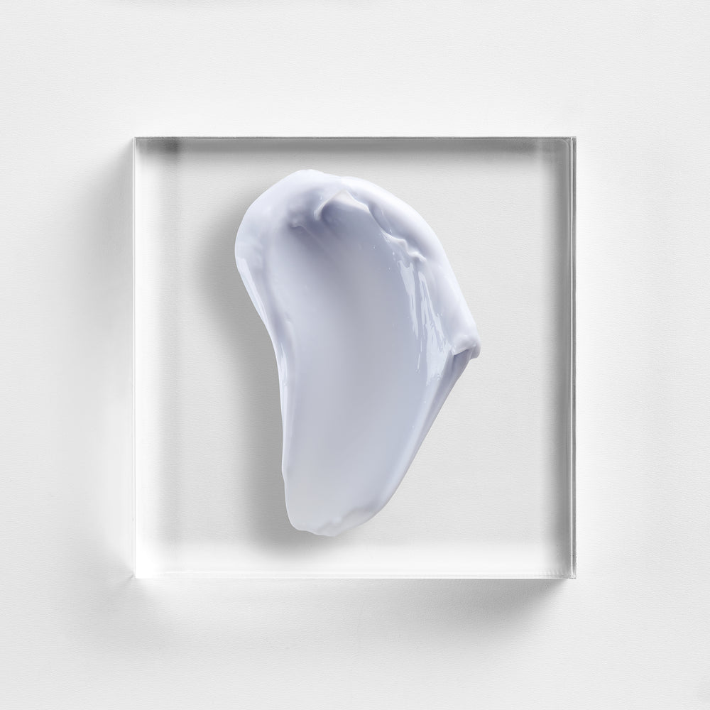 A square clear acrylic block displaying a thick, textured smear of pale lavender-gray cream or gel, set against a clean white background. Close-up of NEOVA Crème de la Copper cream texture, rich moisturiser for smoother, firmer, more hydrated-looking skin.