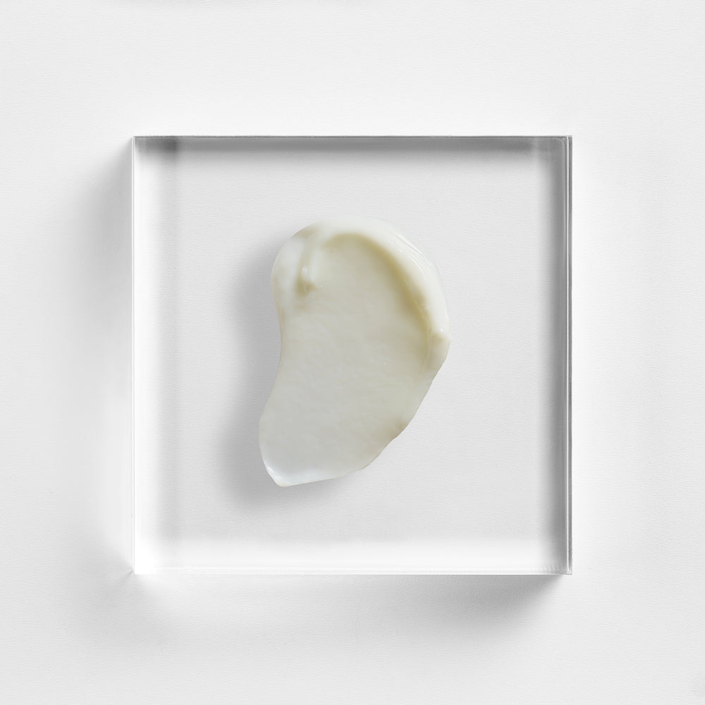 A small irregular cream-colored product sample, likely a cosmetic cream or balm, displayed inside a clear square acrylic block on a clean white background. Close-up texture of NEOVA Overnight Repair, night cream with glycolic acid, lactic acid and salicylic acid.