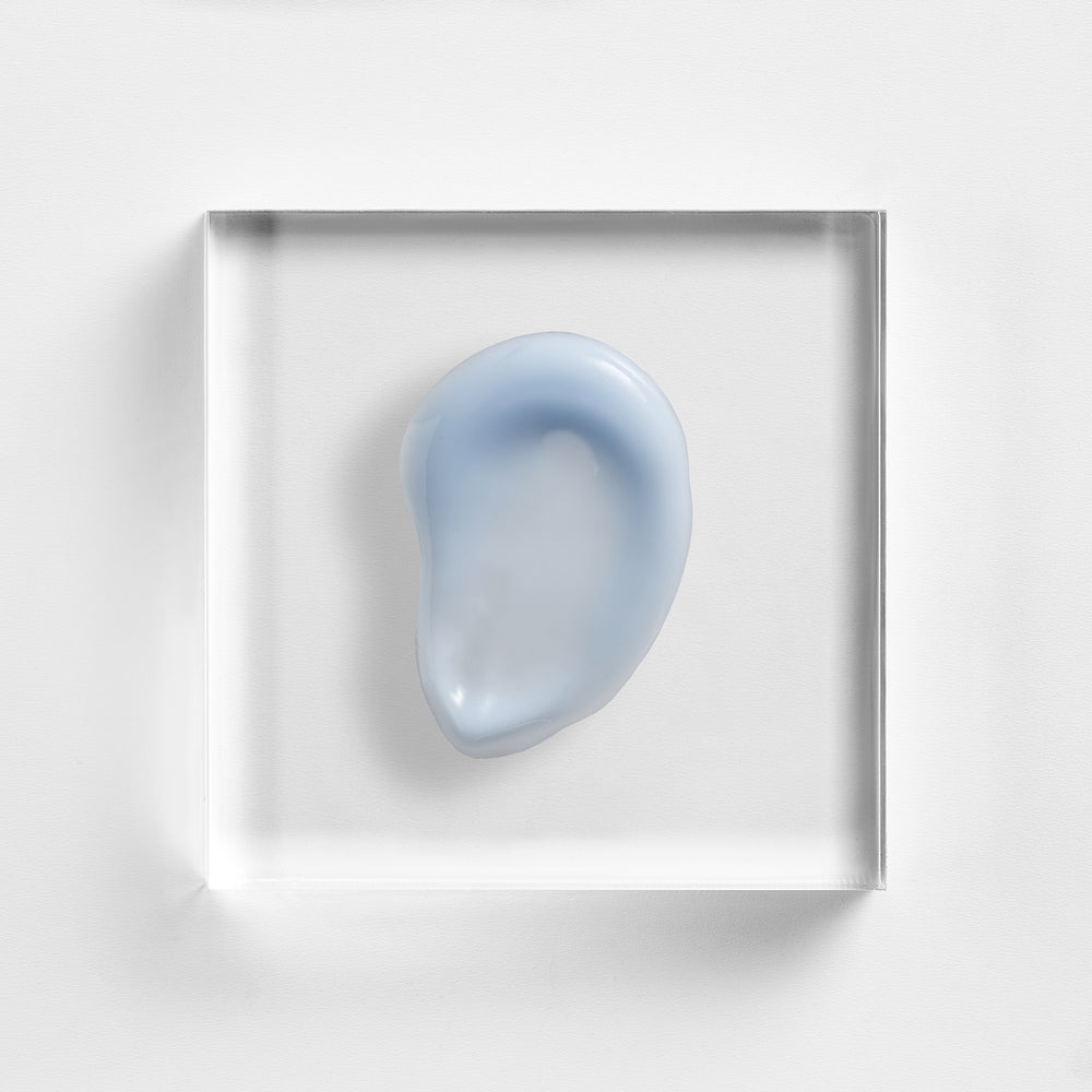 A light blue, organic-shaped translucent object displayed inside a clear square acrylic block on a clean white background. Close-up of NEOVA Super Eye Lift Serum gel texture, designed to smooth fine lines and refresh tired-looking eyes.