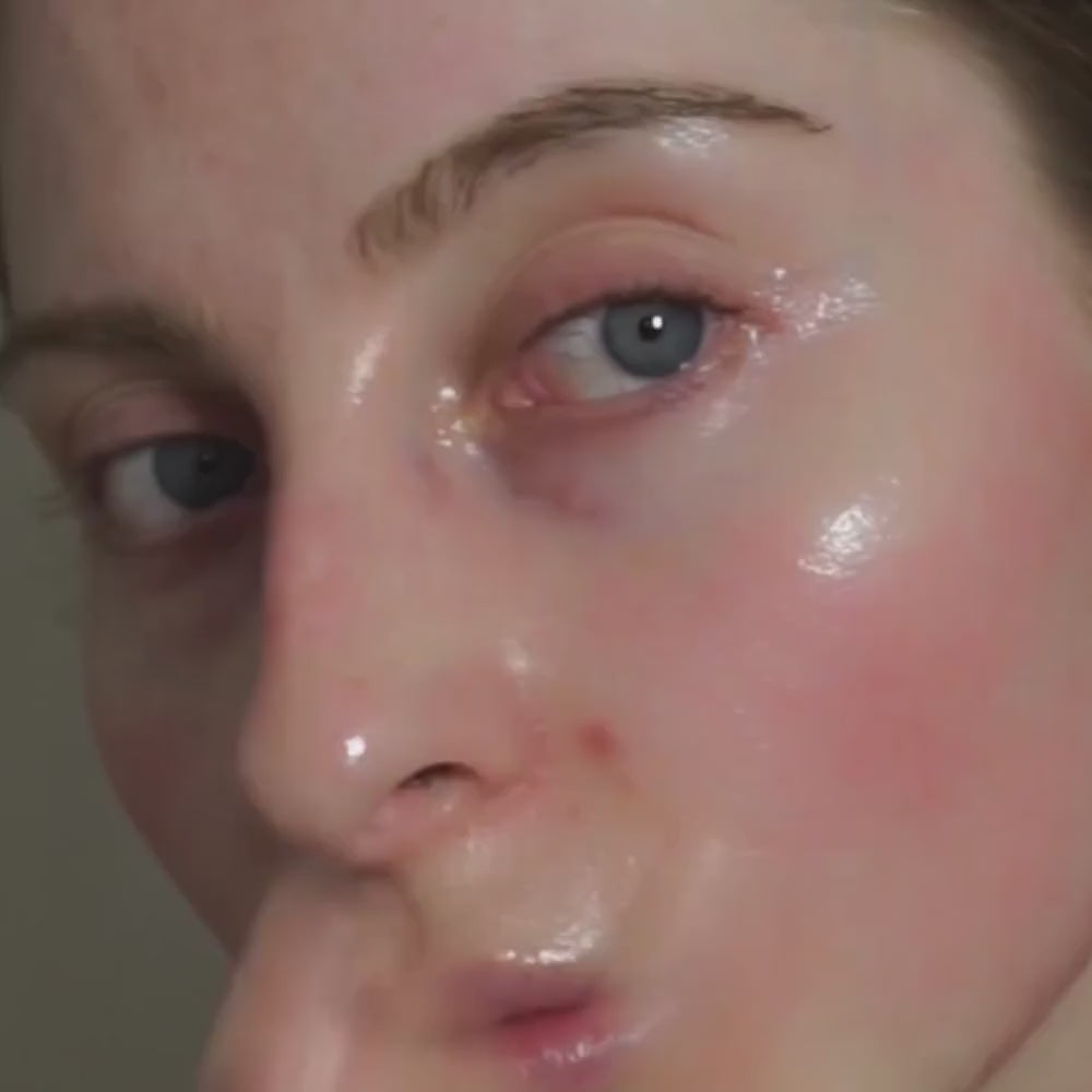 Extreme close-up of a person's face with a glossy, dewy skincare finish on the skin, showing blue eyes, a defined eyebrow, and a pinkish flush on the cheek against a neutral background. Decree Peptide Emollient Veil+ moisturiser applied to the face and neck as an evening skincare step.