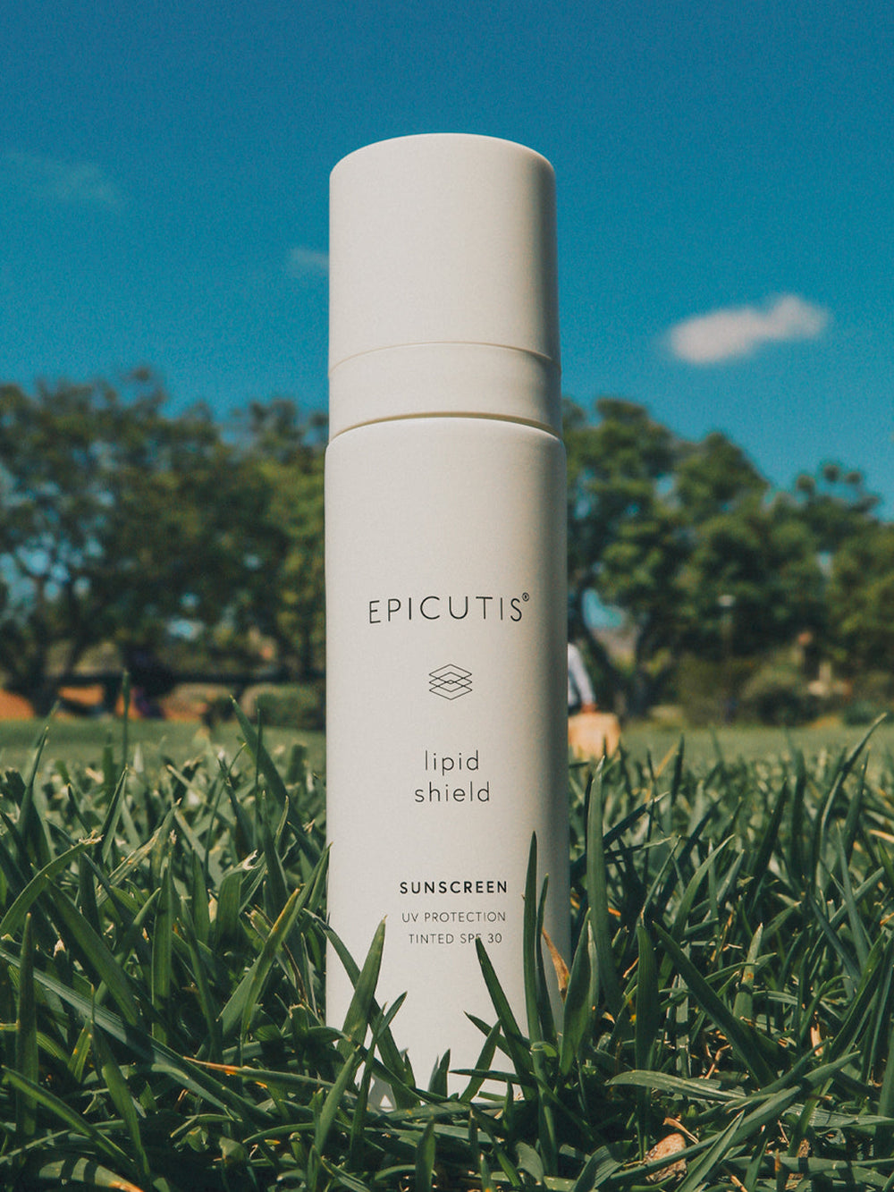 Epicutis lipid shield sunscreen bottle on grass with trees and blue sky in the background