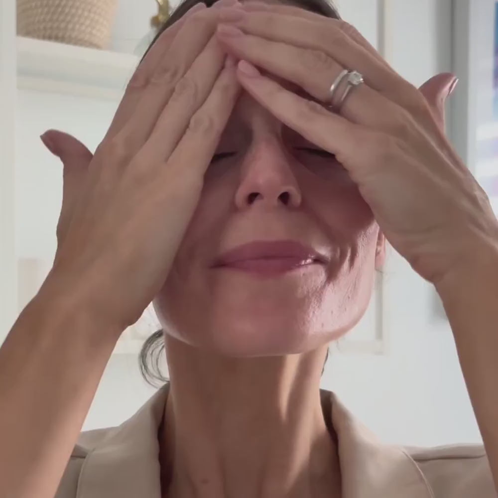 Decree Peptide Emollient Veil moisturiser applied to the face and neck as the final skincare step. A close-up shot of a woman with her eyes closed, holding her hands to her forehead. She is wearing a beige top and a silver ring on her finger, set against a bright, softly blurred indoor background.