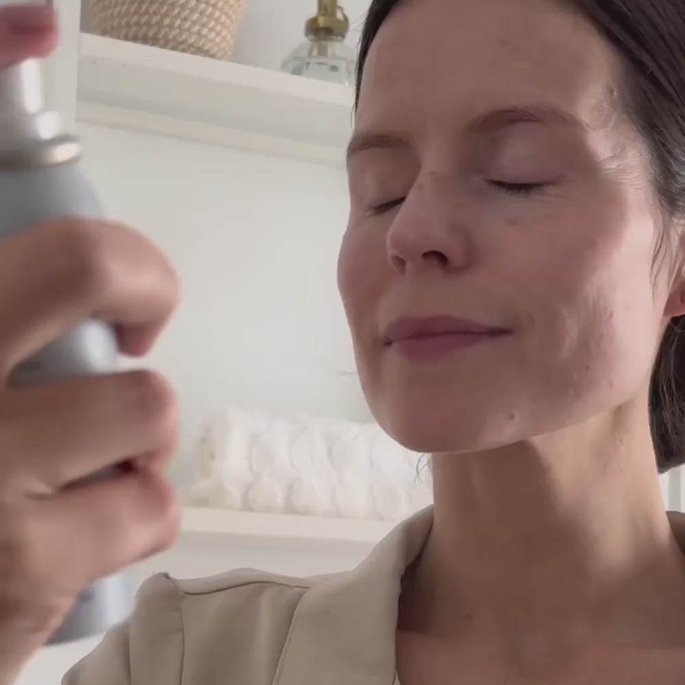 A close-up lifestyle shot of a woman with her eyes closed, holding a silver spray bottle near her face, in a bright, modern room with white shelves in the background. Decree Preparatory Mist sprayed onto the face and neck as the first step after cleansing.