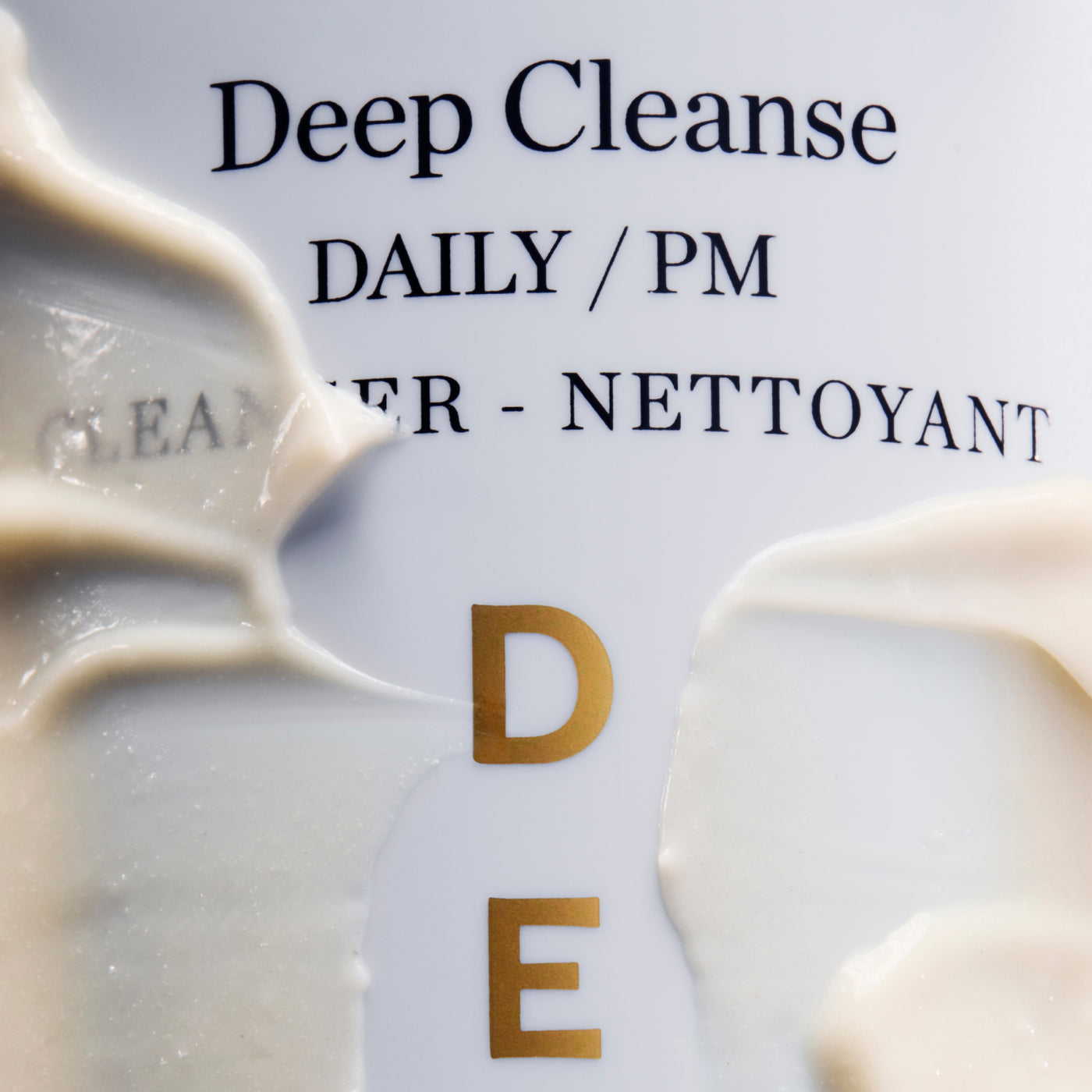 Close-up of a skincare product label reading 'Deep Cleanse DAILY / PM' with cream-colored product swatches smeared across the front and gold letters spelling 'DE'.