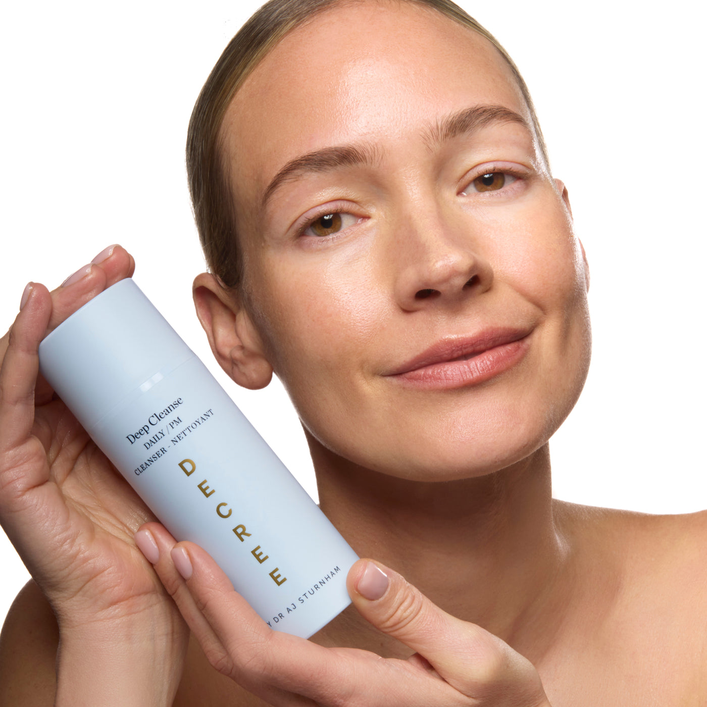 A woman holding a light blue cylindrical skincare bottle labeled 'Deep Cleansing Balm' against a clean white background. Decree Deep Cleanse texture with purifying clay and exfoliating acids for smoother-looking skin.