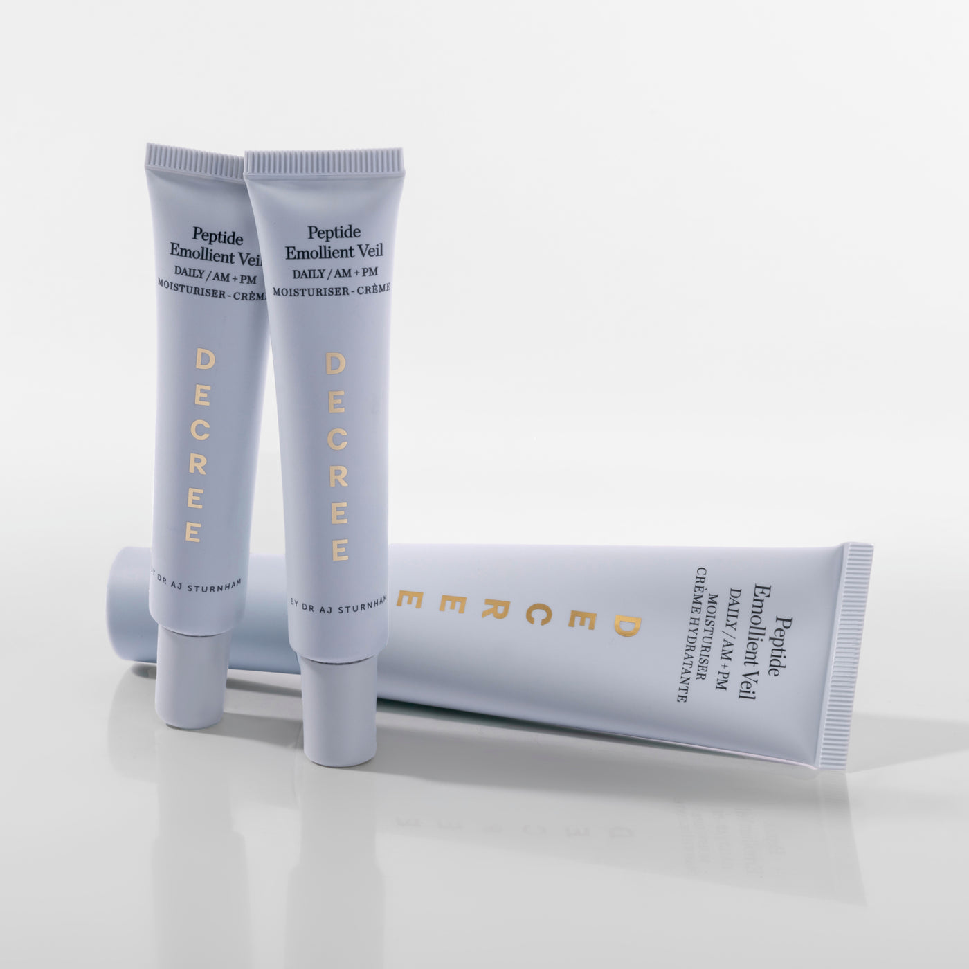 Decree Peptide Emollient Veil 50 ml packaging for firmer, smoother, and more radiant-looking skin. Two light blue cosmetic emollient gel tubes standing upright with one lying horizontally behind them on a clean white studio background. The tubes are branded DECREE and feature gold lettering.