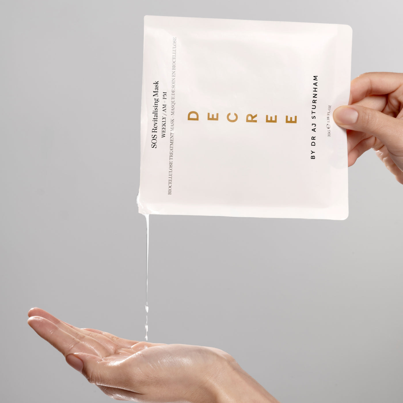 Hand holding a Decree skincare mask packet with a clear sheet being applied to the palm on a gray background. Decree SOS Revitalising Mask essence texture enriched with rose water, glycerin, aloe vera, and panthenol.