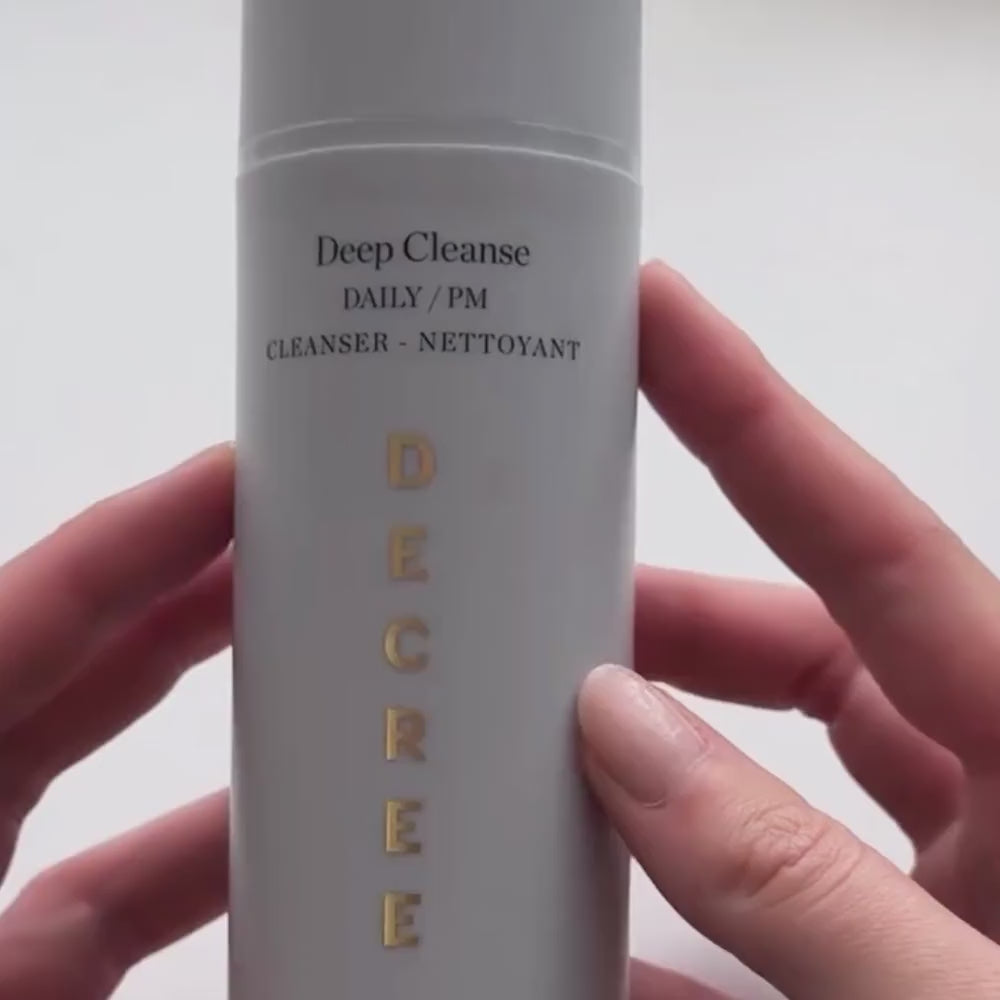 A close-up of a hand holding a white cylindrical skincare bottle labeled 'Deep Cleanse' with vertical gold 'DECRE' lettering, against a plain light background. Decree Deep Cleanse cleanser packaging for congested, oily, combination, and blemish-prone skin.