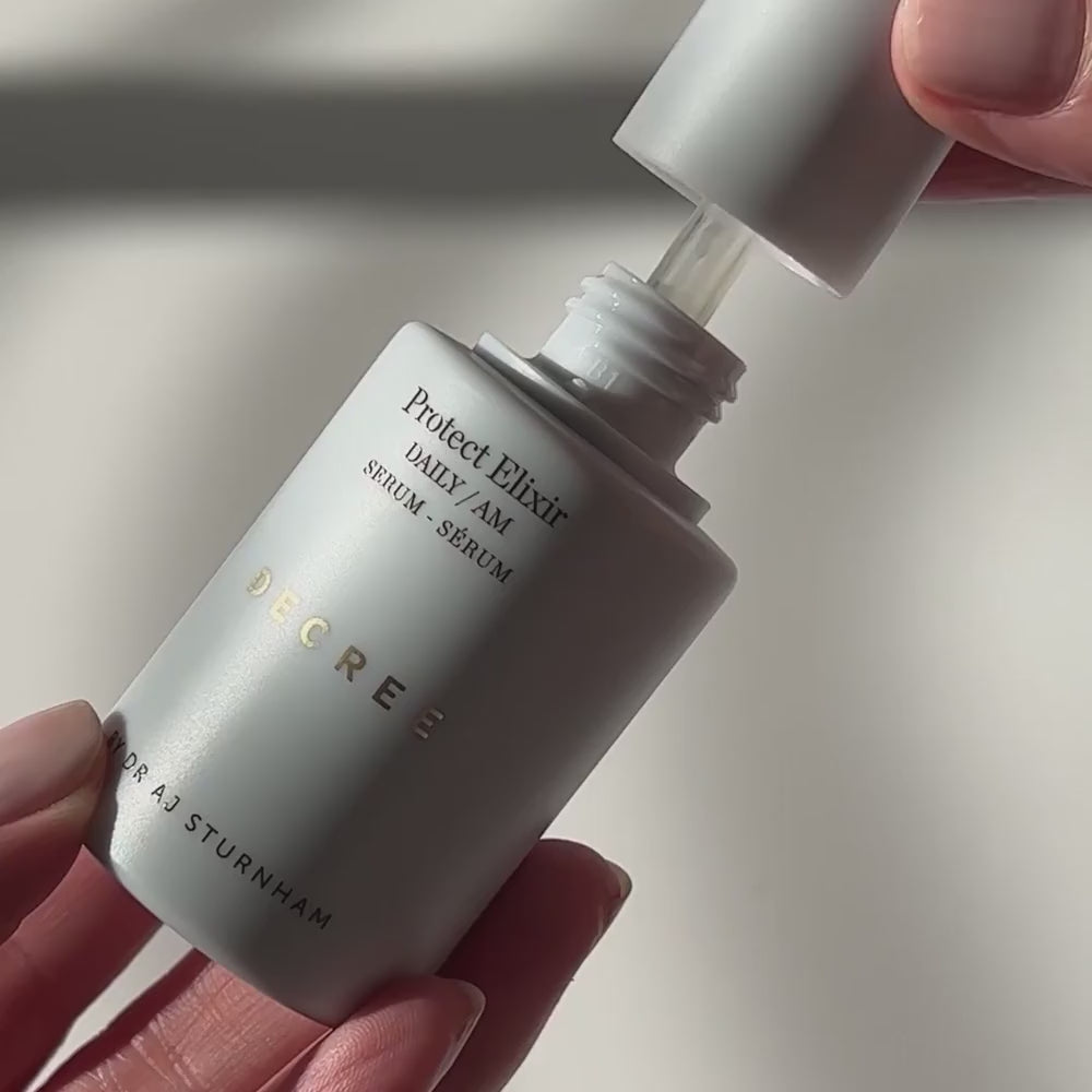 A close-up of a hand holding a frosted white skincare bottle labeled 'Protect Elixir Daily AM Serum' by Decree, with the cap being removed by another hand against a softly lit neutral background.