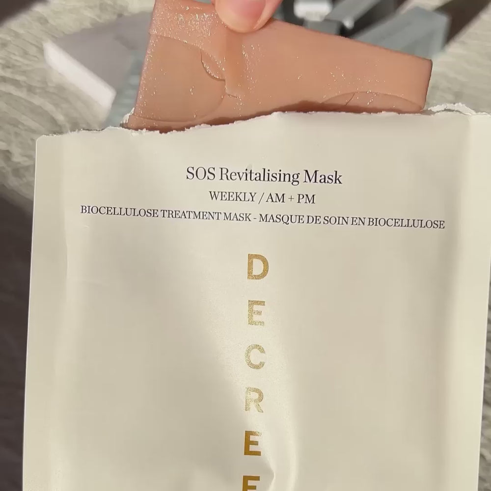 A hand is holding a peach-colored biocellulose mask sheet above its off-white paper packaging, which features gold vertical lettering spelling 'DECREE'.